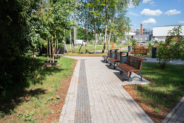 serene urban park scene with wooden benches along a paved pathway under bright blue sky, surrounded by lush green foliage and distant modern buildings on a sunny day, blue, peaceful, fresh, open