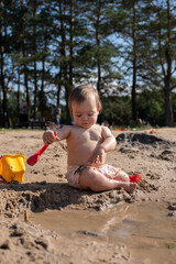 a young child plays intently with a red shovel and yellow bucket, exploring wet sand and a shallow puddle in a sunlit forested area, with trees providing a natural, peaceful backdrop, sandplay, grow
