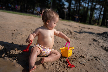 playful toddler enjoys sunny day in muddy sandbox with yellow bucket and red rake amidst green park...