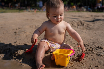 a toddler plays joyfully in a sunny sandy area, scooping and exploring with a small yellow bucket and red scoop, surrounded by the warmth and relaxation of a busy day outdoors, leisure, peaceful