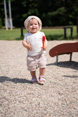 adorable toddler wearing sunhat and sandals joyfully plays outside with a red toy spoon in hand at...