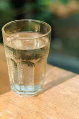 A close-up of a clear glass filled with cold water was placed on a wooden surface. The glass shows condensation droplets on its exterior, indicating the water is chilled. 