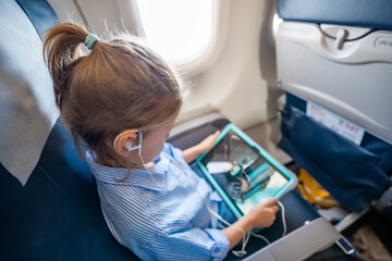 Little girl watching a cartoon on a tablet with headphones during an airplane flight. Digital entertainment, child focus, and quiet family travel experience.