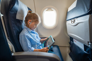 Little girl watching a cartoon on a tablet with headphones during an airplane flight. Digital entertainment, child focus, and quiet family travel experience.