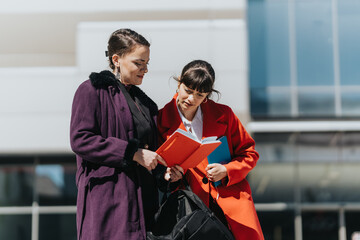 Two businesswomen exchange ideas outdoors, reviewing notes and documents collaboratively. Their...