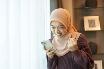 Muslim woman in hijab celebrating good news with smartphone excited and smiling checking messages,...