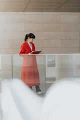 A focused businesswoman dressed in a vibrant red coat holds a notebook while standing in a modern...