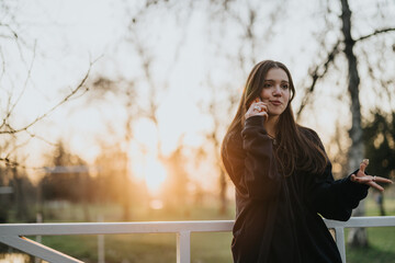 A girl in a dark outfit, standing on a bridge during sunset, talking on her phone and enjoying the...