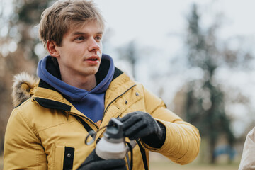 Young man in a yellow jacket and gloves standing outside on a chilly autumn day, holding a water...