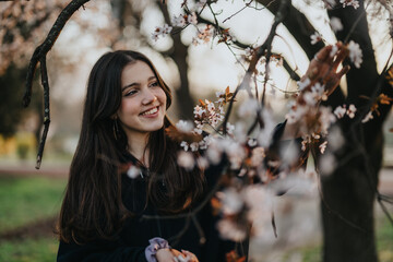 Girl smiling while gently touching a blossoming branch in a serene park during springtime. The...