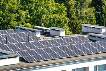 Modern solar panels on a flat building roof with ventilation systems, surrounded by lush green trees. Clean energy and eco-friendly technology in an urban setting © Maciej Bonk