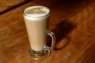 Freshly brewed cup of hot, foamy cappuccino with decorative milk froth topping in a tall, glass on a wood-grain table.