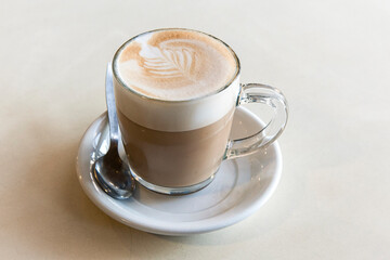 Freshly brewed cup of hot, foamy cappuccino with decorative milk froth leaf topping in a glass cup on a white background.