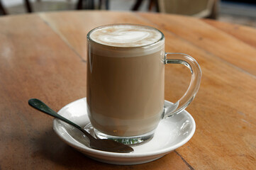 Freshly brewed cup of hot, foamy cappuccino with decorative milk froth topping in a tall, glass mug on a wood-grain table.