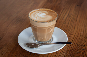 Freshly brewed cup of hot, foamy cappuccino with decorative milk froth topping in a glass cup on a wood-grain table.