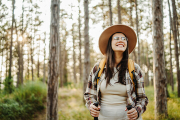 Young caucasian woman hiking or trekking through scenic forest with hiking sticks during golden hour