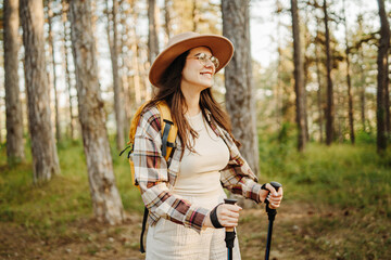 Young caucasian woman hiking or trekking through scenic forest with hiking sticks during golden hour
