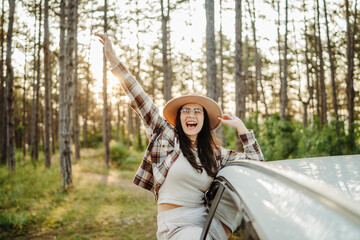 Young caucasian woman sitting on car window in the forest enjoying nature