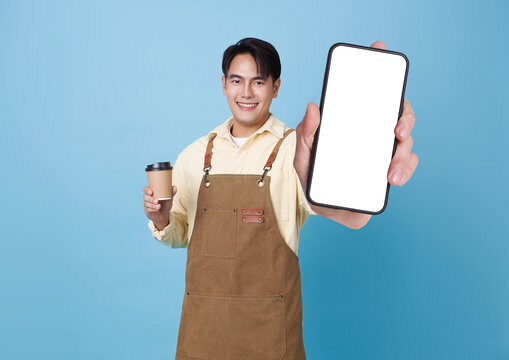 smiling Asian man in apron holds coffee cup and presents smartphone with blank white screen ideal for showcasing coffee shop apps or promotions