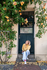 Young woman sitting on doorstep under blooming rose bush in front of vintage house facade