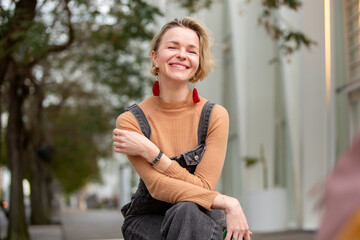 woman sits casually outdoors, smiling warmly at the camera. Her relaxed body language and confident...