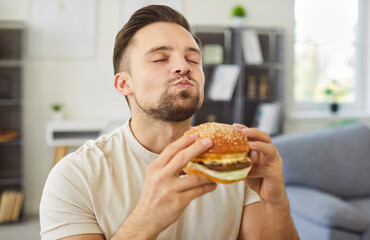 Satisfied young man enjoying delicious burger at home, closing eyes with pleasure and chewing slowly. Portrait of pleased hungry guy appreciating taste of fast food and experiencing food delight.