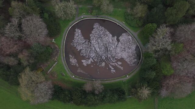 Aerial view of a murky pond surrounded by a circular stone border and lush green trees, creating a contrast of textures and tones, Peebles, Scotland, United Kingdom.
