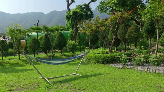 A striped hammock stretches across a vibrant green lawn surrounded by manicured trees and garden beds at Glamtree Resort in Gapyeong, South Korea, with mountains in the background.