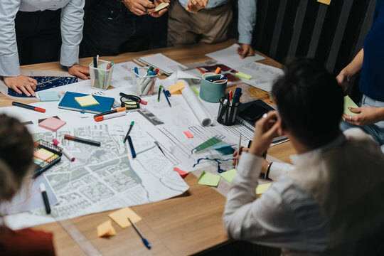 A multiracial business team collaborates on project solutions during a late evening brainstorming session. They analyze plans and share ideas around a conference table filled with maps and stationery. - Powered by Adobe