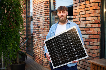 Portrait of bearded man holds solar panel, standing in front of brick building and lush greenery....
