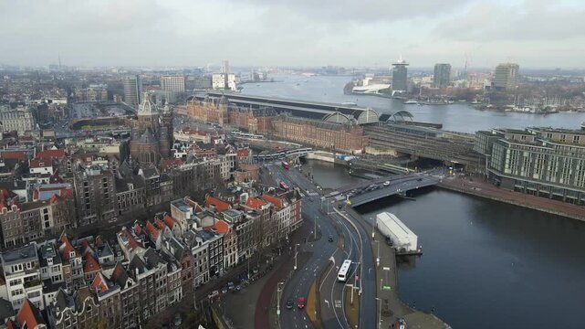 Aerial view of the Amsterdam Centraal Station, with its red brick facade, nestled alongside the IJ river under a muted sky, Amsterdam, North Holland, Netherlands.