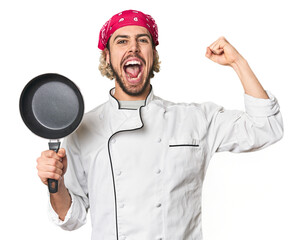 Young Caucasian male chef with frying pan raising fist after a victory, winner concept.