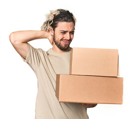 Young man with cardboard boxes in studio touching back of head, thinking and making a choice.