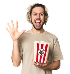 Young man holding popcorn for a movie smiling cheerful showing number five with fingers.
