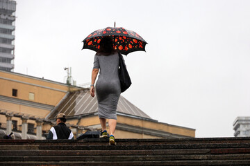 Woman wearing grey summer fitted dress walking up the steps with umbrella on city buildings...