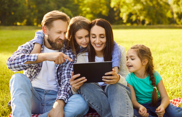 Portrait of happy cheerful young family with children sitting on green grass using tablet in the park. Smiling parents enjoying weekend together with kids in nature browsing internet outdoors.