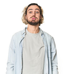 Young Caucasian man in studio setting blows cheeks, has tired expression. Facial expression concept.
