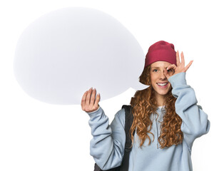 Redhead holding a comic speech bubble excited keeping ok gesture on eye.