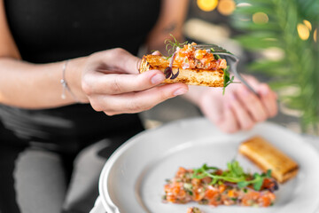 Close-up of a hand holding toasted bread topped with diced vegetables and herbs, served on a white plate with a fork. Gourmet snack or appetizer in elegant presentation.