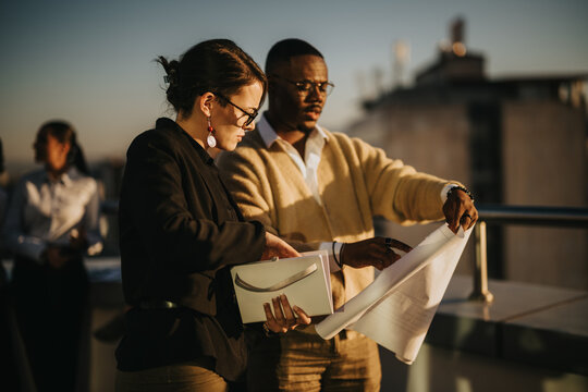 A group of diverse business colleagues work together on a high tower balcony at sunset, collaborating on documents and projects. The vibrant setting enhances teamwork and innovative discussions.