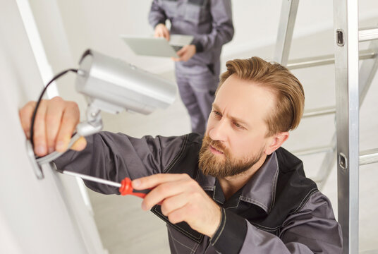 Young technician man in uniform installing surveillance CCTV camera on the wall indoors. Male worker using screwdriver while fixing security camera in modern office or at home. Safety concept.