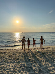 Four happy children holding hands and running into the sea at sunset on a sandy beach. Joyful summer moment, symbolizing freedom, friendship, and carefree childhood.