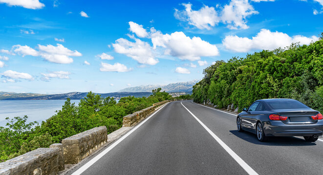 A black car drives along a scenic coastal road with a bright blue sky and distant mountains.