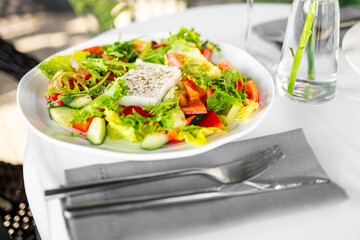 Fresh vegetable salad with feta cheese, served on a white plate with cutlery and water glass on table. Top view, healthy meal concept, colorful and appetizing.