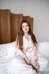 Young woman relaxes in a cozy bedroom wearing pajamas while sitting on a bed surrounded by soft white bedding