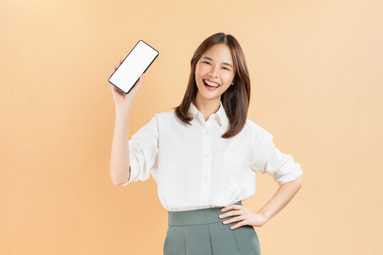 Confident Asian businesswoman holding smartphone mockup of blank screen and smiling on beige color background.