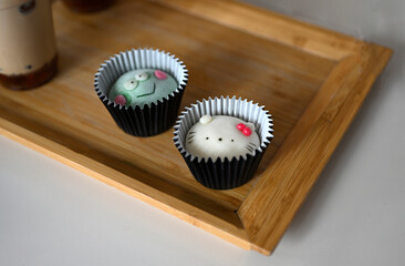 A close-up image of three adorable mochi or steamed rice cakes characters arranged in black cupcake liners on a wooden tray with bubble tea in plastic cups with straws 
