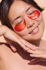 Asian woman with glowing skin, wearing red eye patches, is relaxing with a serene expression,...