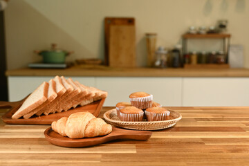 A plate of croissants and muffins sits on a wooden table. The table is surrounded by various kitchen items, including a knife, a spoon, and a bowl. The scene conveys a cozy and inviting atmosphere