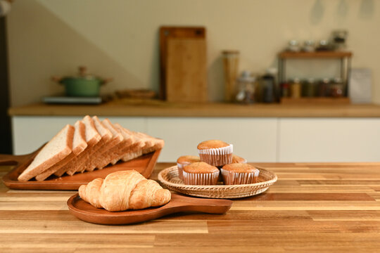 A plate of croissants and muffins sits on a wooden table. The table is surrounded by various kitchen items, including a knife, a spoon, and a bowl. The scene conveys a cozy and inviting atmosphere